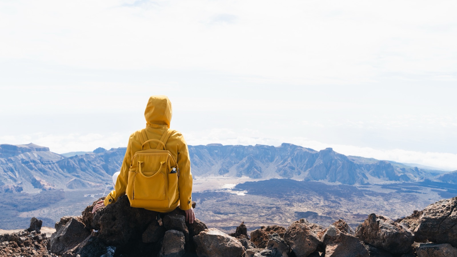 Hiker in a yellow jacket resting on lava rocks high on Mount Teide, overlooking the caldera