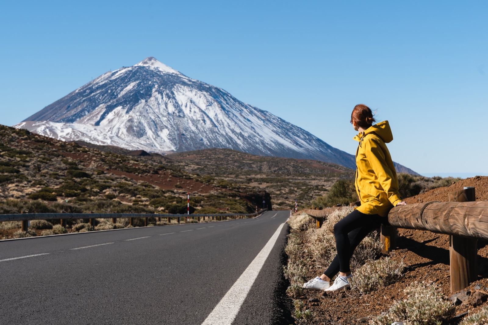 Traveller sitting on a wooden barrier beside the park road with snow-capped Mount Teide ahead